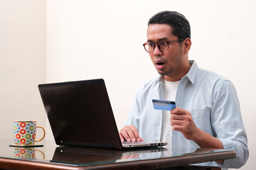 Wall Mural - A man looking to his laptop showing shocked expression while holding credit card