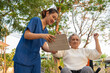 © suwanb - Young nurse and elderly asian senior woman on wheelchair, Asian careful caregiver teach patient do stretches to exercise arm mucsel with digital tablet at the garden on the evening. Health insurance.