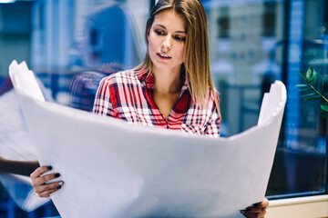 Professional young architect concentrated on reading blueprint of project construction working overtime in office,skilled female student holding big paper with graphic drawing prepare hometask