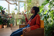 Satisfied relaxed African American woman using smartphone sitting on cozy armchair surround by indoor plants. Pleased black female rest with mobile phone surfing social media in cozy home garden. 