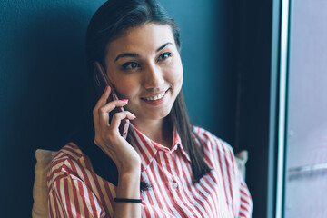 Closeup image of beautiful cheerful female blogger dressed in stylish shirt having mobile conversation via cellular.Cute smiling hipster girl talking on modern smartphone while sitting indoors