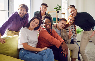 Wall Mural - Portrait of a group of happy young diverse friends men and women sitting on sofa in the living room at home looking cheerful at camera enjoying meeting. Friendship, party and home leisure concept.