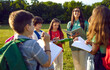 © Studio Romantic - Group of schoolchildrens with their young female teacher hold lessons in nature on sunny day. Junior students and teacher talk standing in park in circle with pencils and notebooks.