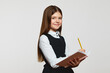 Cheerful elementary schoolgirl in uniform holding open notebook and looking at camera, standing isolated over white background