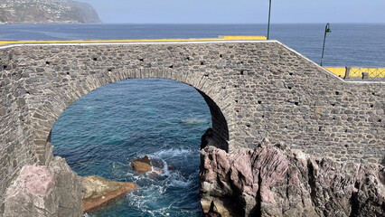 Wall Mural - A stone arch bridge overlooks the rocky shore and turquoise waters of the ocean, with waves crashing below, set against a distant coastal landscape.