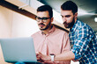 © BullRun - Skilled male teacher of it school explaining programming tools to student learning on course, hipster guy helping his friend for updating software on laptop computer working in office together
