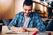 © BullRun - Smiling male blogger satisfied with having great idea for publication making notes sitting at desktop with netbook, positive student enjoying learning process doing homework task in college library