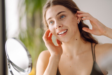 Wall Mural - Beautiful woman looking in the mirror, touching her face with her hand, examines acne, pigmentation. Medicine and cosmetology.