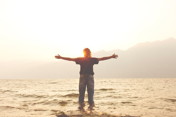 woman with open arms welcomes the warmth of the light of the setting sun on the banks of a lake