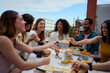© CarlosBarquero - Group of multi-ethnic young friends enjoying meal gathered on rooftop. Happy millennial people having fun drinking red wine and eating celebrating a party event on summer day