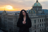 Fototapeta Zwierzęta - Attractive young curly brunette woman in a black jacket without lingerie sits on a rooftop against a historic building during a cozy sunset