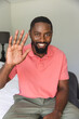 African American man waving during a video call, sitting on bed at home
