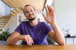 Caucasian young man sitting at table, waving hello at home on a video call