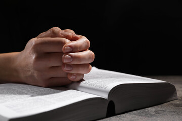 Wall Mural - Religion. Christian woman praying over Bible at table against black background, closeup. Space for text