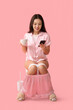 Young Asian woman in pajamas with coffee cup and mobile phone sitting on toilet bowl against pink background