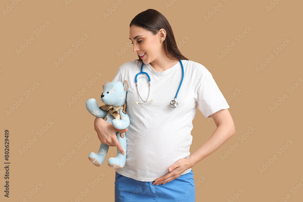 Beautiful young pregnant doctor with teddy bear on brown background