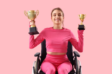 Poster - Sporty young woman in wheelchair with gold cups on pink background