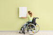 Young woman in wheelchair hanging painting on green wall, back view