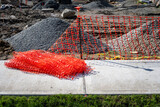 Fototapeta Pomosty - Fresh new orange safety fencing being set up next to sidewalk around a construction site of dirt and gravel piles
