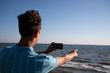 © vladorlov - A young attractive guy is posing while relaxing on the sea and taking a photo with his phone.