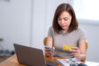 © Wasana - A woman is sitting at a desk with a laptop and a cell phone. She is looking at her cell phone and holding a credit card. Concept of modern technology and convenience