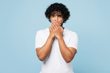 Wall Mural - Young shocked surprised astonished sad Indian man he wears white t-shirt casual clothes cover mouth with hands isolated on plain pastel light blue cyan background studio portrait. Lifestyle concept.