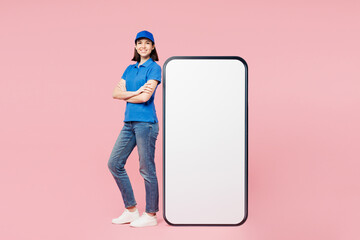 Wall Mural - Full body delivery employee woman wears blue cap t-shirt uniform workwear work as dealer courier stand near big huge blank screen mobile cell phone with empty area isolated on plain pink background.