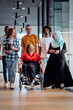 © .shock - A diverse group of young business people walking a corridor in the glass-enclosed office of a modern startup, including a person in a wheelchair and a woman wearing a hijab