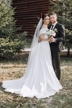 A bride and groom are posing for a picture in front of a log cabin. The bride is wearing a white dress and the groom is wearing a black suit. They are holding a bouquet
