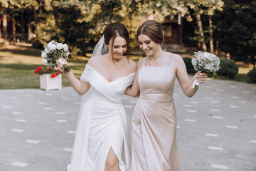 Wall Mural - Two women are walking together, one of them is wearing a white dress and the other is wearing a tan dress. They are both holding bouquets of flowers