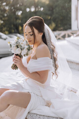 Wall Mural - A woman in a white dress sits on the ground holding a bouquet of flowers. She is wearing a veil and a necklace. The scene is serene and peaceful, with the woman looking at the camera