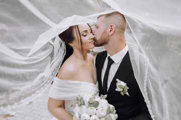 Wall Mural - A bride and groom are kissing under a white veil. The bride is holding a bouquet of flowers