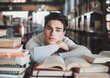 Young Man Studying Intently Among Stacks of Books in Library, Academic Dedication and Focus