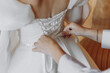 A woman is adjusting the straps of a wedding dress. The dress is white and has a lace belt. The woman is wearing a bracelet and a ring. The scene is intimate