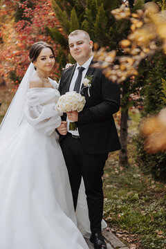 A bride and groom are posing for a picture in a park. The bride is wearing a white dress and the groom is wearing a black suit. They are holding a bouquet and a vase, respectively