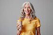 Smiling elderly American woman in yellow t-shirt with glass of protein drink in hand on gray background. Food concept for adult food products, silver economy.