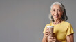 Smiling older Latino woman in yellow t-shirt with glass of protein drink in hand on gray background. Food concept for adult food products, silver economy.