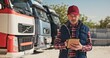 © VAKSMANV - Handsome mature Caucasian man wearing red cap standing next to trucks while using tablet device. Hardworking driver typing message or text for delivery order. Logistics company concept.