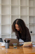© Wasana - A woman is sitting at a desk with a laptop and a notebook. She is looking at the laptop and she is in a serious or focused mood