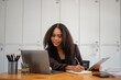 © Wasana - A woman is sitting at a desk with a laptop and a notebook. She is smiling and she is working on something. The scene suggests a positive and productive atmosphere