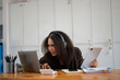 © Wasana - A woman is working on her laptop and tablet, with a look of frustration on her face. She is surrounded by various office supplies, including a calculator, a pen, and a cup