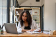 © Wasana - A woman is sitting at a desk with a laptop and a stack of papers. She is looking at the papers and seems to be focused on her work. Concept of productivity and concentration