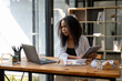 © Wasana - A woman is sitting at a desk with a laptop and tablet in front of her. She is wearing a white shirt and black tank top. The room has a wooden desk and a bookshelf with many books
