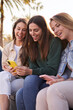 Vertical. Group of generation z beautiful female friends smiling sitting on street using and looking mobiles phones. Addiction of technology and social networks of young happy people sunny day outdoor