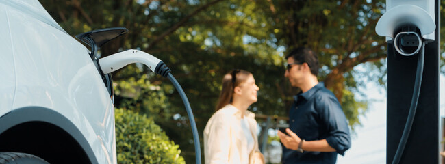 young couple recharge electric car battery from charging station in green city park in springtime. r