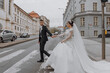 A bride and groom are walking down a street, with the bride wearing a long white dress. The man is holding the woman's hand, and they are both smiling. The scene is a romantic
