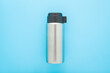 Closed new silver steel thermos with dark black plastic mug for hot drink or soup on light blue table background. Pastel color. Closeup. Top down view.