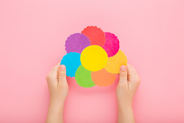 Wall Mural - Baby girl hands holding and showing beautiful flower head shape from colorful paper on pink table background. Pastel color. Closeup. Point of view shot. Little child creating gift. Top down view.