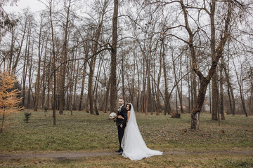 Wall Mural - A couple is posing for a picture in a forest. The bride is wearing a white dress and the groom is wearing a suit. Scene is romantic and peaceful