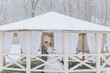 A couple is kissing in the snow under a pavilion. The pavilion is covered in white fabric and has a snow-covered roof. The scene is romantic and intimate